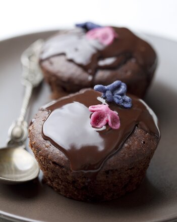 Foto von zwei Bananen-Kakao-Muffins auf einem Teller. Die Muffins sind mit Schokolade übergossen und mit Blüten garniert. | © SONNENTOR