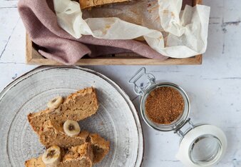 Auf dem Foto ist ein Bananen Zimt Brot mit Bananen garniert zu sehen. Davor liegen auf einem Teller aufgeschnittene Brotscheiben. | © SONNENTOR