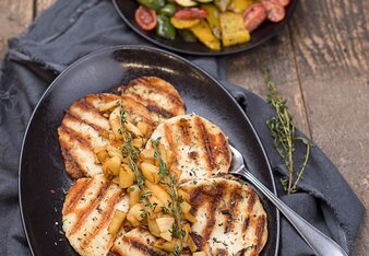 Auf dem Foto ist ein schwarzer Teller mit Grillkäse und Apfelchutney darauf zu sehen. Im Hintergrund sieht man ein Teller mit buntem Grillgemüse. | © SONNENTOR