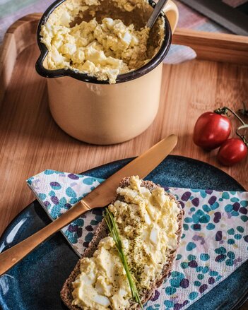 Auf dem Foto ist eine Scheibe Brot mit Eiaufstrich zu sehen. Im Hintergrund sieht man ein ganzes Gefäß mit Aufstrich. | © SONNENTOR