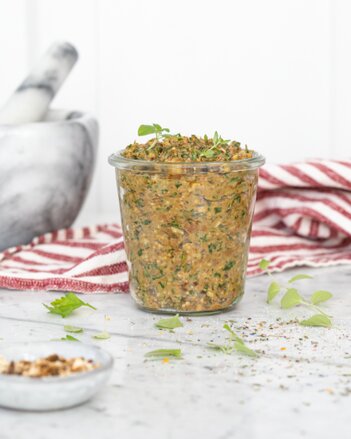 Auf dem Foto sieht man ein Glas mit Steinpilz-Pesto. Daneben sind verschiedene Kräuter zu sehen. Im Hintergrund sieht man einen Mörser. | © SONNENTOR