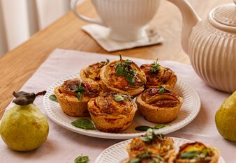 Die Mini Erdäpfel Quiches liegen auf 2 Tellern verteilt, dekoriert mit Basilikum und Majoran Blättern. Im Hintergrund steht eine Tasse Tee, 2 Birnen liegen daneben.  | © SONNENTOR