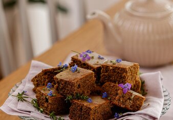 Die Schwarzteekuchen Stückchen liegen auf einer Etagere und sind mit kleinen blauen und violetten Blümchen dekoriert. Im Hintergrund steht eine Teekanne.  | © SONNENTOR