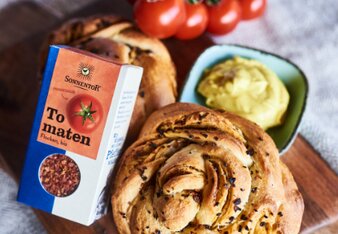 Auf dem Foto sieht man zwei Stück Tomaten-Swirl-Bread. Danben sieht man eine Schüssel mit Curry-Linsen-Dip. Auch eine Packung Tomatenflocken ist zu sehen. | © SONNENTOR