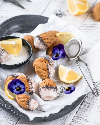 Auf dem Foto sind mehrere Zitronen-Mohn-Madeleines zu sehen. Daneben sieht man Blüten. | © SONNENTOR