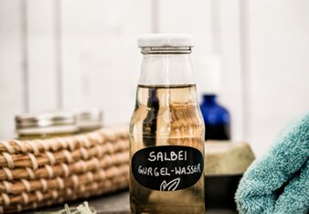 Auf dem Foto ist eine Flasche mit Salbei-Gurgelwasser zu sehen. Daneben sieht man Salbei und auch ein Handtuch ist auf dem Foto zu sehen. | © SONNENTOR