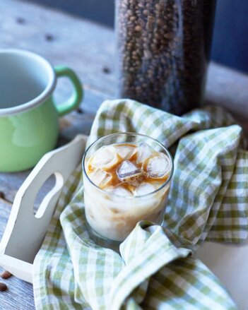 Auf dem Foto ist ein Glas mit Chai Eiskaffee zu sehen. Das Glas steht auf einem grün kariertem Küchentuch. Dahinter sind Kaffeebohnen zu sehen. | © SONNENTOR