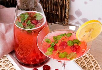 Ein Cocktailglas steht neben dem Krug, beides gefüllt mit einem roten Mocktail, gemacht aus Himbeeren und dem SONNENTOR Tschin Tschin Sirup. Kleine Minzblättchen schwimmen darin, das Glas ist mit einer Zitronenscheibe garniert. | © SONNENTOR