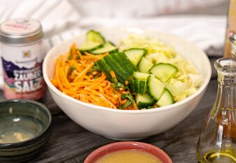 Die Salatschüssel ist gefüllt mit Karottenstiften, Gurkenstücken und Weißkraut und steht neben einer Dose Ayurvedischen Zaubersalz, einem Schälchen mit Dressing und einem Holzlöffel mit Salz. | © SONNENTOR