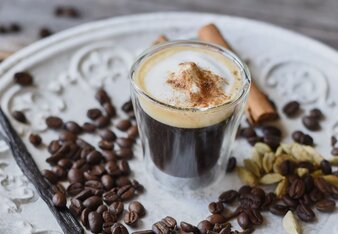 Auf dem Foto sieh man ein Glas mit einem Espresso macchiato darin. Daneben liegen Kaffeebohnen, Zimtstangen und Vanilleschoten. | © SONNENTOR