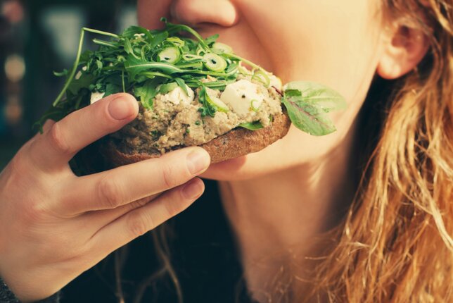 Auf dem Foto sieht man ein Brot mit Aufstrich und frischen Kräutern darauf. Eine Frau beißt gerade von dem Brot ab. | © SONNENTOR